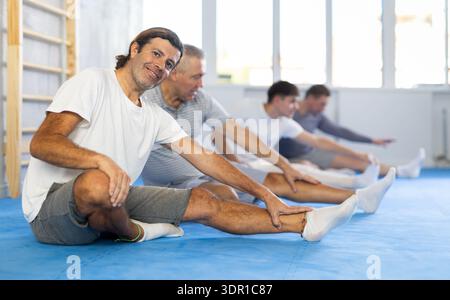 Hommes en vêtements de sport à l'entraînement de groupe assis sur le sol et étirant les muscles Banque D'Images
