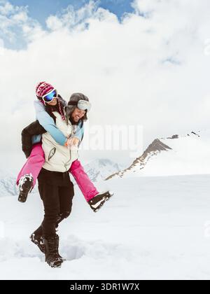 Joyeux jeune couple marche piggyback dans la neige profonde partager le lien romantique sur la montagne enneigée, symbolisant l'amour, le bonheur, la liberté, la relation ludique, Banque D'Images