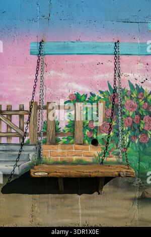 Banc en pierre saillant soutenu par des chaînes métalliques contre une murale peinte vibrante avec des roses, une clôture en bois et un fond pastel. Banque D'Images