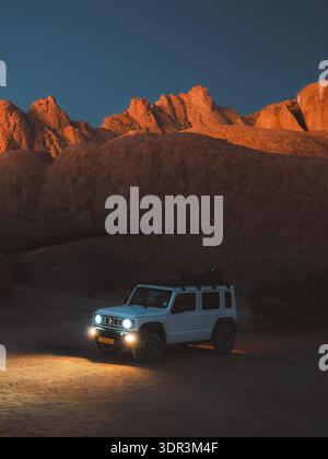 Vue d'un SUV blanc stationné sur un sol sablonneux avec ses phares allumés, sur fond de montagnes accidentées et ensoleillées sous un ciel crépusculaire, Spitzkoppe, Banque D'Images