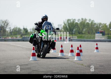 Un motocycliste sur une moto d'aventure verte naviguant à travers un parcours de slalom de cônes rouges et blancs sur un terrain d'entraînement pavé. Banque D'Images