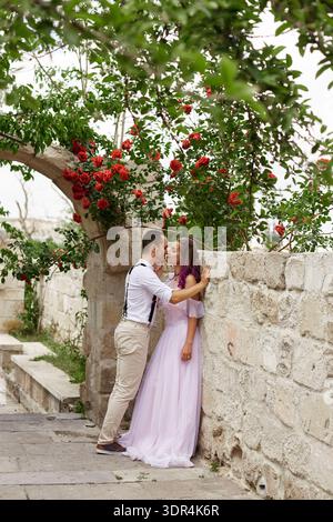couple profite d'un moment ensemble dans un jardin, debout près d'un mur de pierre. Ils se penchent pour un baiser au milieu des fleurs en fleurs, capturées à la lumière du jour. Banque D'Images