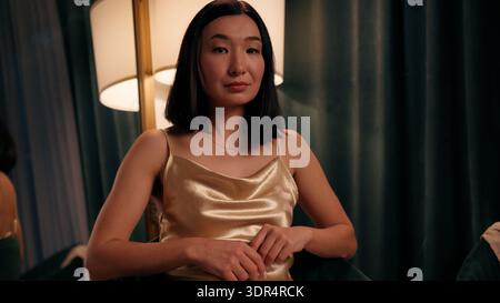 Portrait calme femme asiatique sérieuse assise à la maison soir gros plan japonaise coréenne chinoise femme à l'intérieur regardant l'appareil photo confiant portant doré Banque D'Images