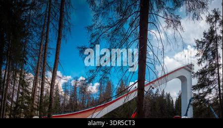 Rampe de saut à ski s'élevant au-dessus de la forêt alpine à Cortina d Ampezzo, en Italie, avec une structure en béton spectaculaire, un ciel bleu, la lumière de l'hiver et des pins imposants Banque D'Images
