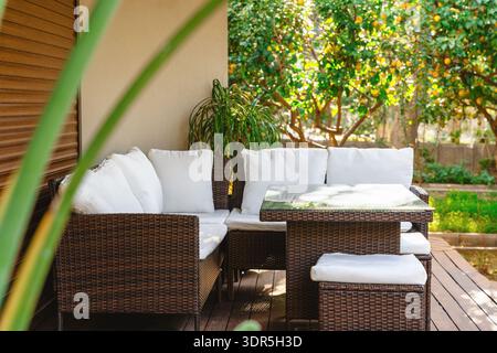 Canapé extérieur en osier et table sur une terrasse ombragée à côté d'un jardin luxuriant, invitant à la détente et aux repas dans la lumière du soleil Banque D'Images