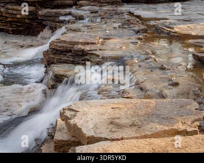 Eau en cascade sur des roches calcaires stratifiées avec de la glace fondante au début du dégel printanier en lumière douce du soleil Banque D'Images
