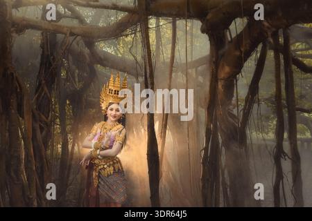 Danseuse apsara thaïlandaise souriante en vêtements traditionnels et coiffe (mokot) dansant dans un paysage forestier mystique, Thaïlande Banque D'Images