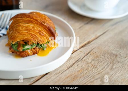 Gros plan d'un croissant savoureux gourmand rempli de légumes verts et d'un œuf poché sur une table en bois avec une tasse de café Banque D'Images