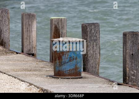Une borne bleue rouillée se dresse sur une jetée en béton entourée de poteaux en bois altérés avec l'eau ondulée en arrière-plan Banque D'Images