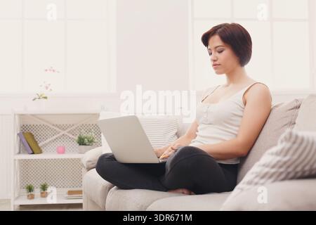 Femme travaillant sur ordinateur portable dans le salon tout en étant assis confortablement sur canapé Banque D'Images