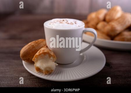 tasse de cappuccino mousseux en porcelaine blanche saupoudrée de cacao à côté d'une profiterole fourrée de crème mordue sur une soucoupe striée, table chaude en bois avec Banque D'Images