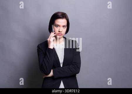 Femme parlant au téléphone tout en portant costume noir dans le studio avec fond gris Banque D'Images