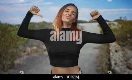 Femme avec l'estomac exposé dans un top de récolte noir sourit et lève ses bras sur un chemin de rue, penché légèrement avec des taches de rousseur visibles ; joie insouciante. Banque D'Images