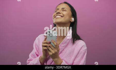 Jeune femme dans un peignoir rose aime la musique avec des écouteurs et un smartphone sur un fond rose vif isolé, exsudant la joie et la détente. Banque D'Images