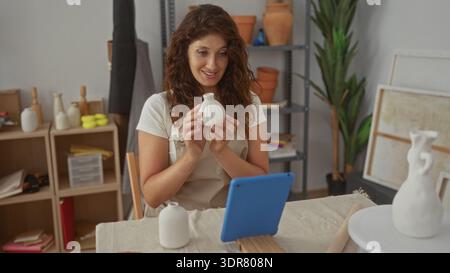 Femme portant tablier tient petit vase en céramique avec les mains et les gestes vers la tablette sur la table de travail en studio ; concentration créative. Banque D'Images