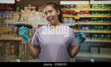 Femme dentiste tient guide de teinte de dent et sondes dentaires, souriant avec les mains gantées dans la construction de supermarché ; confiance ludique. Banque D'Images