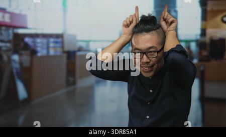 Jeune homme chinois avec des lunettes souriant, les mains sur la tête avec les index vers le haut, portant une chemise marine au terminal de l'aéroport occupé à l'intérieur ; méfait ludique. Banque D'Images