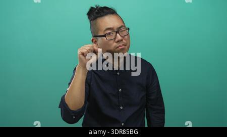 Jeune homme avec la main levée et un petit geste du poing, portant une chemise marine dans un ensemble de studio sarcelle ; scepticisme doute de l'hésitation. Banque D'Images