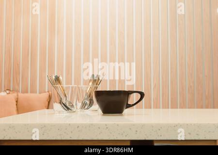 Une tasse à café noire se trouve sur une table blanche à côté d'un ensemble de verres remplis de cuillères en argent. La scène montre une configuration de café simple dans la lumière du matin. Banque D'Images