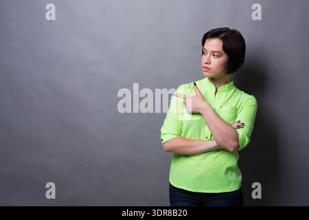 Femme en chemise verte pointe sur le côté sur un fond gris pendant un moment décontracté Banque D'Images