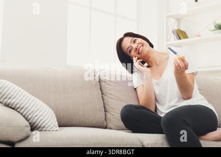 Femme au téléphone sourit tout en étant assise sur le canapé avec test de grossesse en main Banque D'Images