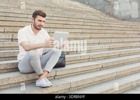 Homme assis sur les marches en utilisant la tablette dans l'espace public pendant la journée dans la ville Banque D'Images