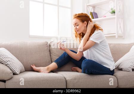 Femme assise sur le canapé regardant le test de grossesse dans sa main avec l'expression réfléchie Banque D'Images