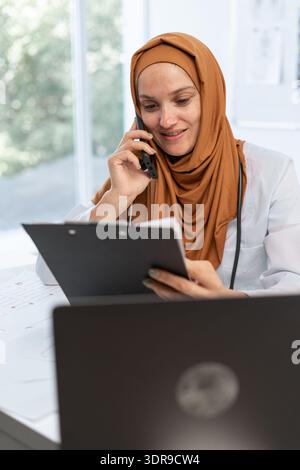 Médecin féminin arabe fournit des conseils médicaux en utilisant le téléphone travaillant dans la clinique moderne. consultations de patients. Banque D'Images