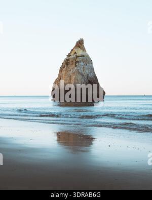 Vue d'une formation rocheuse solitaire se dressant contre les vagues douces, sa texture rugueuse reflétée dans le sable humide, créant un SC côtier serein Banque D'Images