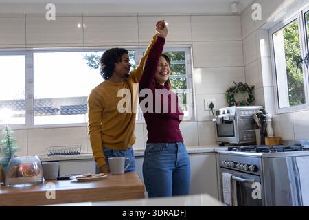 Couple diversifié tourbillonnant dans la cuisine par fenêtre dans des chandails avec tasse et pâtisseries sur le comptoir en bois Banque D'Images