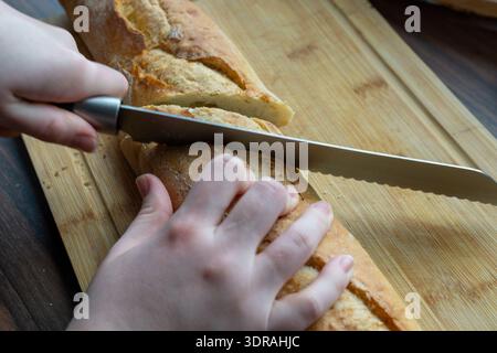 Gros plan d'une personne coupant une baguette avec un couteau dentelé sur une planche de bois. Banque D'Images
