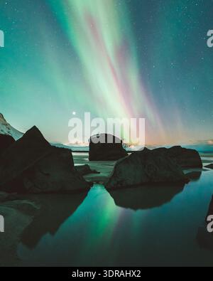Vue sur les danses aurores boréales à travers le ciel nocturne, reflétée dans l'eau calme parmi les rochers sombres et accidentés sur la plage, Ramberg, Nordland, No Banque D'Images