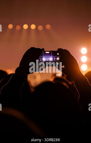 Silhouette d'une personne tenant un téléphone portable pour enregistrer une performance musicale en direct lors d'un festival. La scène est éclairée par des projecteurs ambres chauds cr Banque D'Images