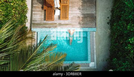Une jeune femme en chapeau et maillot de bain se détache dans l'eau claire d'une piscine privée. Vue de dessus de la magnifique piscine avec lit de bronzage Banque D'Images