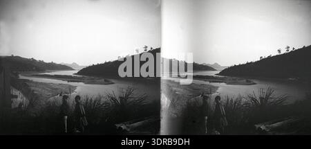 Une photographie stéréoscopique montre un paysage fluvial panoramique en Indochine (aujourd'hui Vietnam) vers 1906-1907. Au premier plan, deux piétons, peut-être d'un groupe minoritaire vietnamien ou chinois basé sur des couvre-têtes, marchent le long de la rive. L'image met en valeur les paysages naturels, les tenues culturelles et l'activité humaine locale au début du XXe siècle en Asie du Sud-est. Banque D'Images