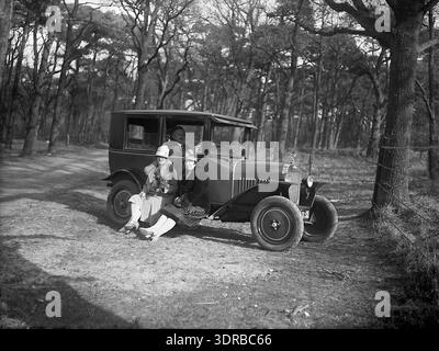 Cette photographie, prise par Jan Stevens entre 1928 et 1932 aux pays-Bas, montre deux femmes posant sur le marchepied d'une petite automobile dans un espace boisé lors d'une sortie de loisir. Le conducteur, assis derrière le volant droit, regarde le photographe. L'image documente le tourisme automobile du début du XXe siècle, l'automobile de loisir, la représentation des sexes, la conception de voitures et les activités de loisirs en plein air dans les paysages néerlandais. Banque D'Images