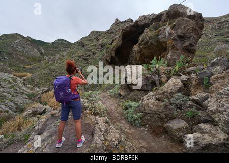 Une femme photographe aux cheveux roux bouclés et un sac à dos violet photographie une grande formation rocheuse volcanique avec une ouverture de grotte proéminente dans la Barra Banque D'Images