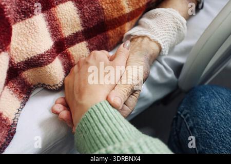 Gros plan d'une femme tenant la main d'un patient âgé sous une couverture. Compassion et accompagnement pendant les traitements hospitaliers et les soins médicaux à l'intérieur. Banque D'Images