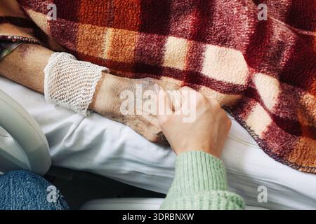 Femme tenant la main d'un patient âgé dans un lit d'hôpital. Accompagnement et soins pendant le traitement médical. Compassion et concept de soins à l'intérieur. Banque D'Images