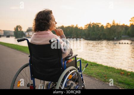 Triste femme âgée assise en fauteuil roulant marchant sur la route en ville en plein air, vue arrière. Vieille dame paralysée dans la chaise pour les personnes avec d. Banque D'Images