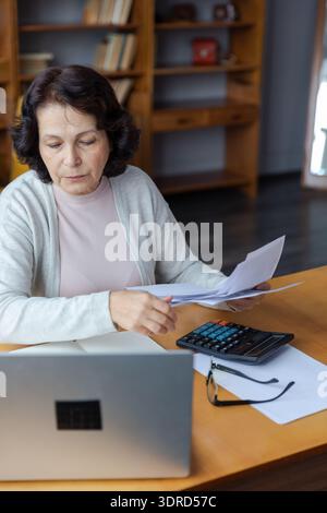 Femme âgée moyenne assise avec un ordinateur portable et un document papier, Pensive vieille dame mature lisant la facture de papier payer en ligne à la maison homme Banque D'Images