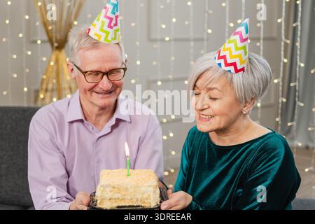 Fais un vœu. Couple senior de famille dans chapeau de fête célébrant l'anniversaire ensemble à la maison. Vieil homme soufflant du burnin Banque D'Images
