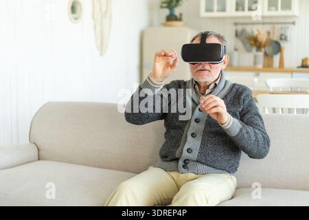 Homme senior mature excité portant à l'aide du casque de lunettes de réalité virtuelle Metaverse VR à la maison. Grand-père touchant l'air pendant la VR Banque D'Images