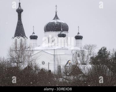 Église orthodoxe russe avec des dômes d'oignon recouverts de neige lors d'une chute de neige hivernale à Vasknarva. Neige tombant sur une église blanche du monastère orthodoxe. Banque D'Images