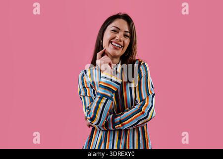Jeune femme heureuse riant et posant dans une chemise rayée colorée sur un fond rose vif, exprimant la joie, la confiance et l'esprit d'énergie dynamique Banque D'Images