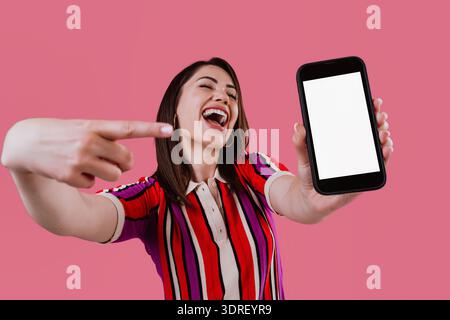 Jeune femme souriante tenant un smartphone avec un écran blanc en avant, présentant une nouvelle application, offre ou concept de communication contre un bac rose vibrant Banque D'Images