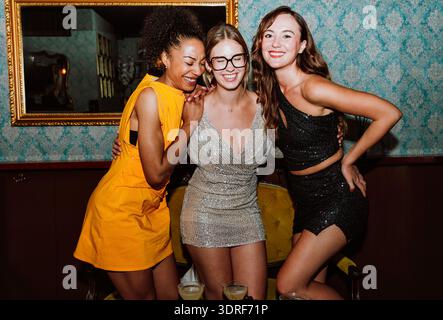 Trois jeunes femmes diverses portant des robes de fête posant ensemble, célébrant des occasions spéciales avec des cocktails et des collations dans un bar élégant ou un salon Banque D'Images