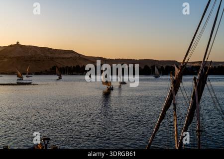 Des felouques silhouettées glissent sur le Nil au coucher du soleil à Assouan, en Égypte. Les lignes épurées et les tons chauds conviennent aux voyages, à la culture et aux visuels de marque. Banque D'Images