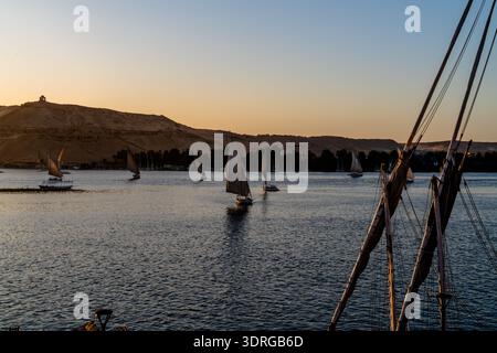 Les bateaux de Felucca naviguent à travers le Nil au crépuscule à Assouan, en Égypte, avec des collines désertiques en arrière-plan. Une scène de rivière calme évoquant la tradition et la lenteur t Banque D'Images