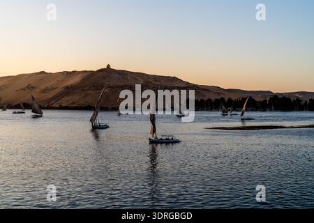 Vue large sur le Nil au coucher du soleil à Assouan, en Égypte, avec de multiples felouques naviguant sur l'eau libre et la lumière chaude du désert. Banque D'Images